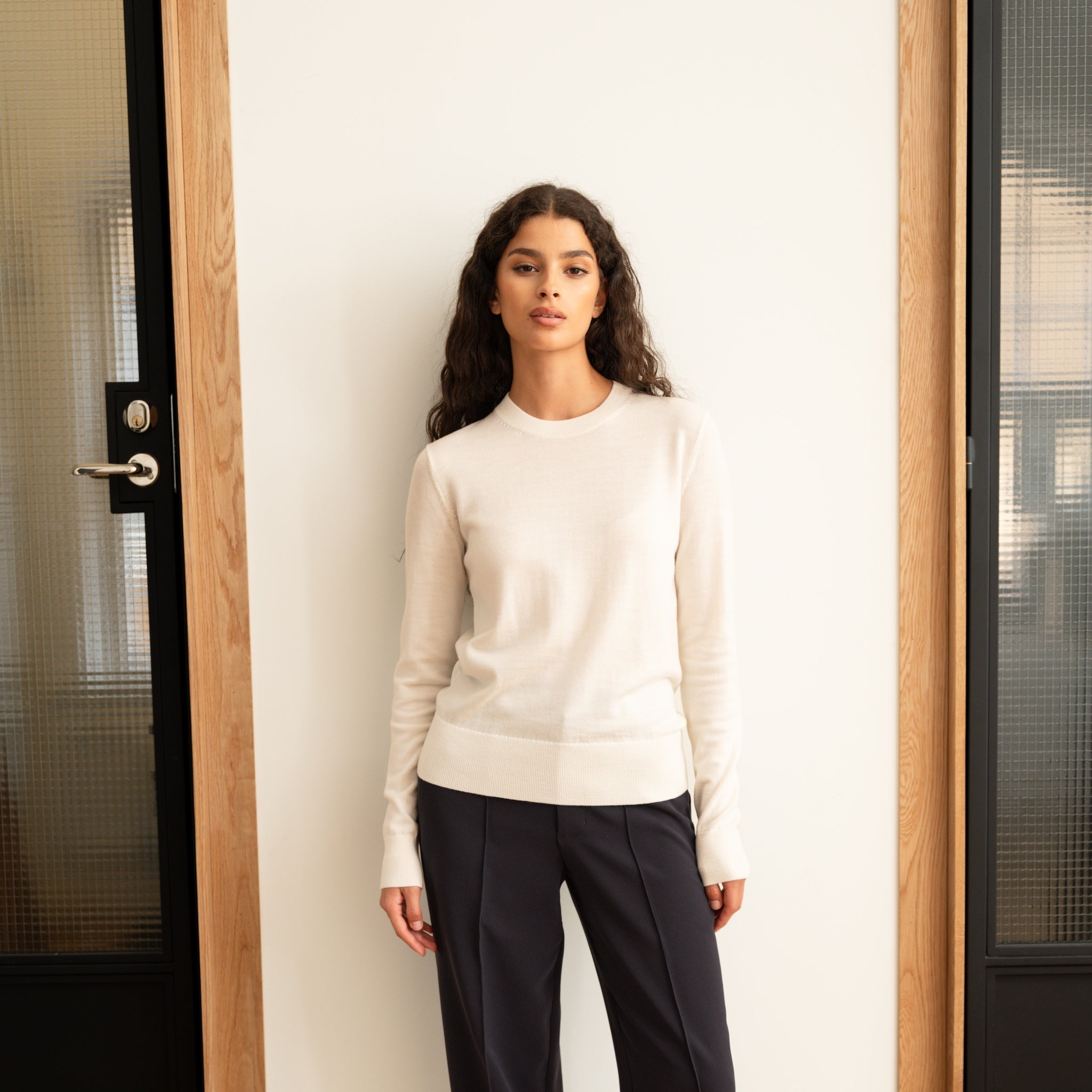 Woman wearing a Ninepine off-white merino wool crewneck sweater with crepe relaxed trousers against a white wall